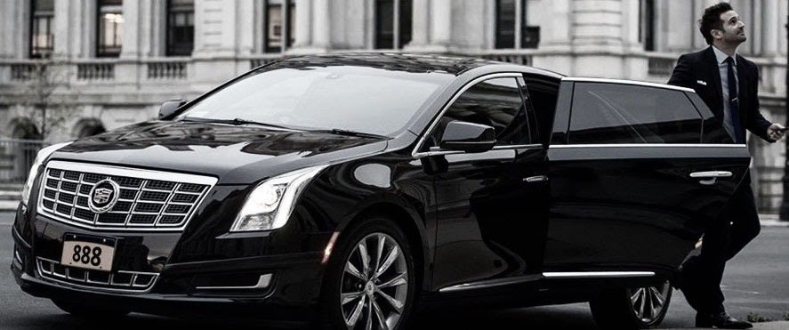 Gateway limousine service's professional chauffeur in a suit standing next to the open rear door of a black Cadillac luxury sedan on a city street in Downtown St. Louis