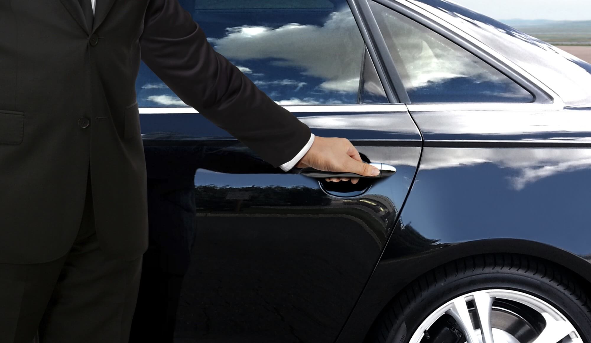 A professional chauffeur in a black suit opening the rear door of a luxury black car for a passenger arriving at St. Louis Lambert International Airport (STL).